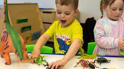 A young boy wearing a yellow shirt with blue dinosaur plays with toy dinosaurs at Science Time