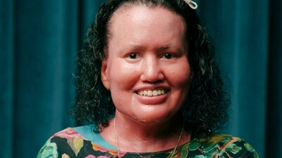 Portrait of Carly Findlay, smiling and wearing a colourful floral top,  in front of a teal curtain