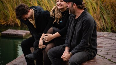 Za Górami © Monika Jakubowska, 2023 ECM Records A young man, blonde woman and bearded man laughing together sitting next to a pond