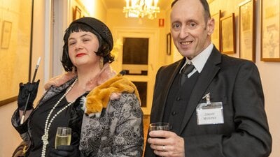 A man and a woman in 1920s dress  stare at the camera. Perhaps they have a secret.