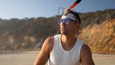 Mitch Tambo sits on a beach with a rocky coastline looking out to sea.