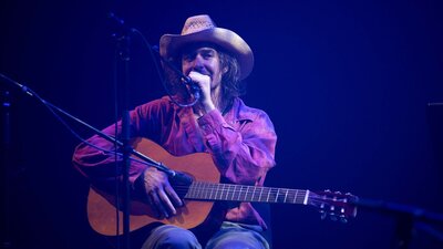 Archer, a man wearing a cowboy hat, performs at a concert. He sits with an acoustic guitar and mic.