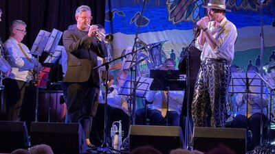 A concert. Archer, in akubra hat, sings into a microphone and Eugene Ball plays trumpet.