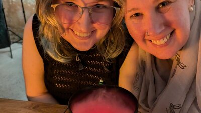 Two women smiling over a cauldron of red, steamy brew