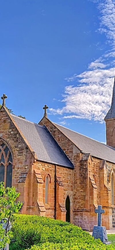 Old graves and gardens surround a sandstone and bluestone church with a spire and bell tower