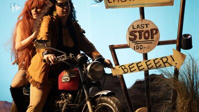 Two women dressed in costumes and wigs on a motorcycle in a theatre like setting