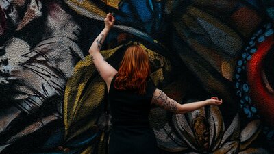 SPHERE A woman with red hair holds her arms away from her. The wall behind is covered in a mural