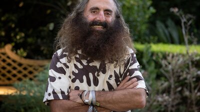 Costa Georgiadis, at the launch of The People’s Portrait in March 2025. Costa Georgiadis stands in a lush garden, smiling. His arms are crossed over a leaf pattern shirt.