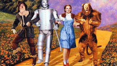 Still from The Wizard of Oz showing a scarecrow, tin man, woman and lion on a yellow brick road