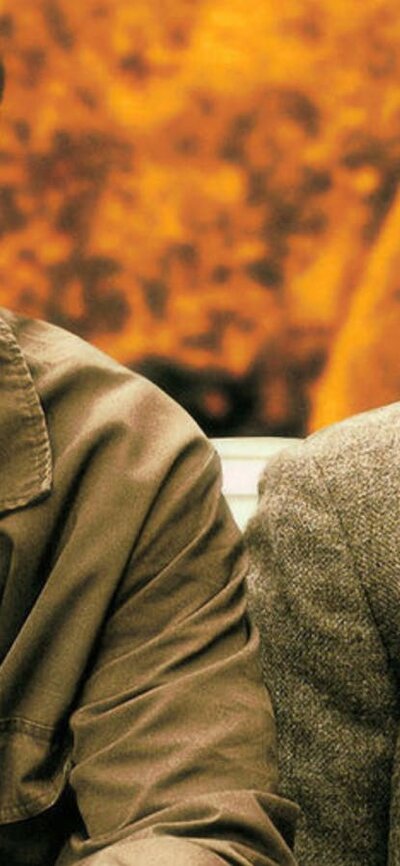 Two men sit next to each other, man on left laughing