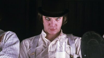 Man wearing hat and make up on right eye glares at camera holding glass of milk