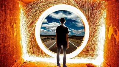 A youth looks through a large round window surrounded by a circle of and strands of light to a road.