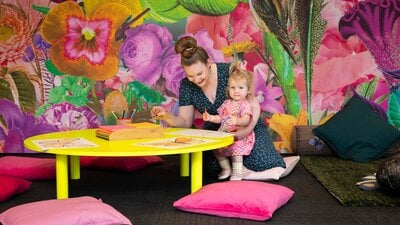 A picture of a woman and a child drawing on a fluorescent yellow table in front of a colourful wall