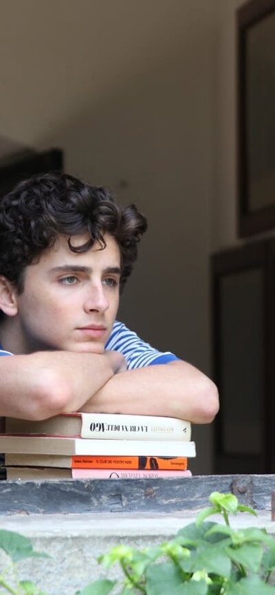 A teenage boy leans on some books and looks out of a window