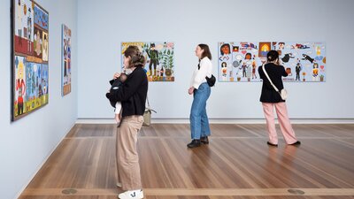 Visitors in Gallery 7 of Super Kaylene Whiskey, looking at Kaylene Whiskey's artwork Three people look at colourful portraits hanging on pale blue walls. One of them holds a baby.