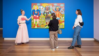 A guide in a pink outfit speaks to two visitors about a colourful portrait.