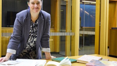 A woman in a grey blazer smiles at the camera while leaning on a wooden table covered with books.