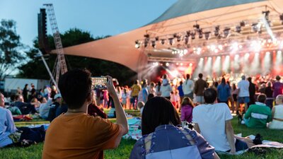 People sitting on grass, looking up at stage as they watch outdoor performance