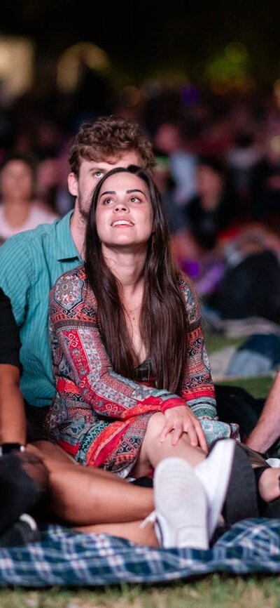 People sitting on grass, looking up at stage as they watch outdoor performance