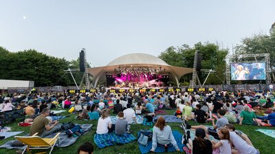 People sitting on grass, looking up at stage as they watch outdoor performance
