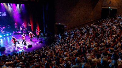 Audience applauds The 60 Four on a well-lit stage in a packed venue.