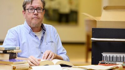 A man wearing glasses and a blue button-down shirt while sitting with books in the National Library