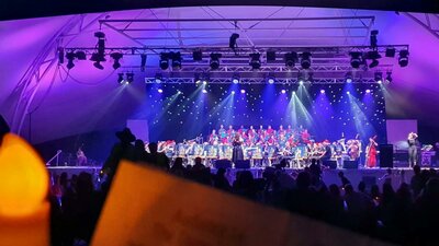 A choir performs on an outdoor stage illuminated by purple stage lights with candles in the audience