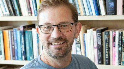 Man with glasses smiles in front of bookshelf