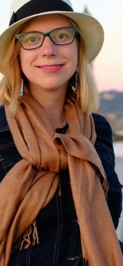 Woman in hat and scarf smiles by waterfront at sunset.