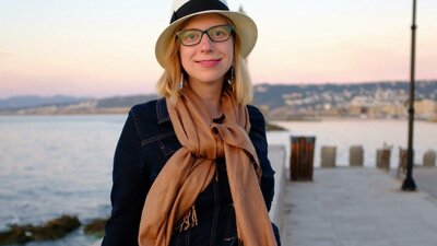 Woman in hat and scarf smiles by waterfront at sunset.