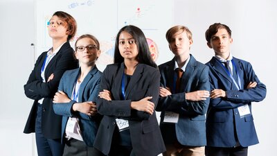 Children wearing suits and lanyards stand with arms crossed, in front of a whiteboard.