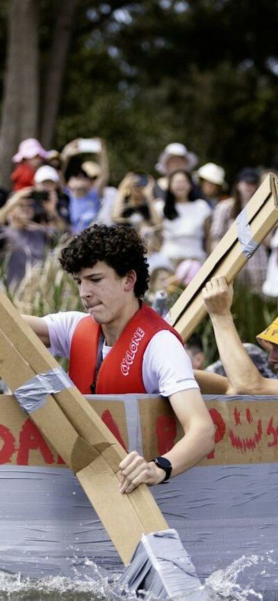Young paddlers racing a cardboard boat on the lake while a crowd watches from the shore
