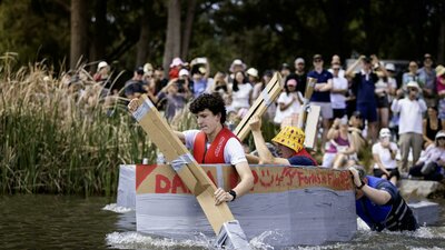Young paddlers racing a cardboard boat on the lake while a crowd watches from the shore