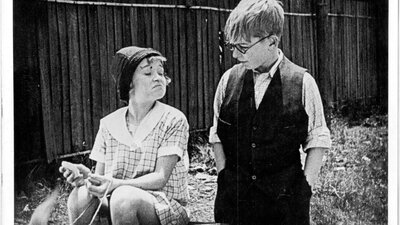 Still from The Kid Stakes (1927) Black and white image of girl smirking and sitting down and boy with glasses looking down at her