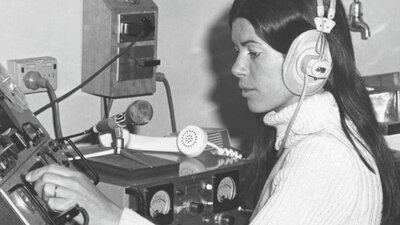 A black and white, vintage photograph of a woman wearing large headphones, intently operating a reel