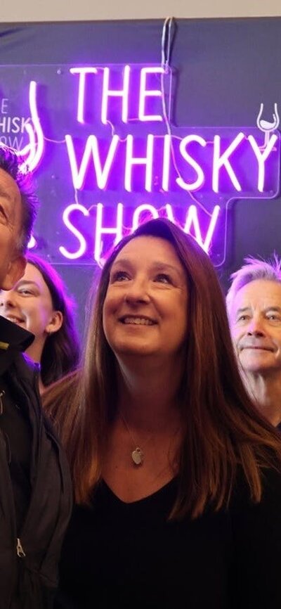 A group gathers in front of a Whisky Show banner and neon sign for a selfie.