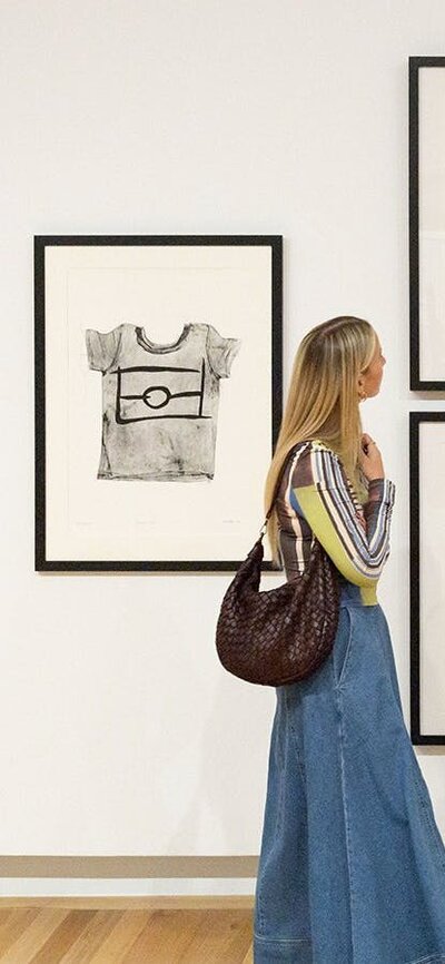 A person holding a brown handbag walks by a Gallery wall, looking at several artworks.