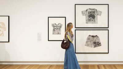 A person holding a brown handbag walks by a Gallery wall, looking at several artworks.