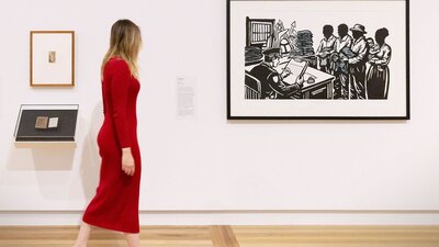 A person wearing a pink dress walks by a Gallery wall, looking at several artworks.