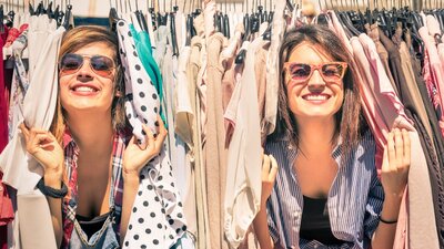 two women smiling peeking through a rack of clothes at an outdoor market