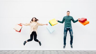 A man and a women holding shopping bags jumping in the air and smiling.
