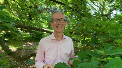 A smiling man with glasses and grey hair standing outside with trees.