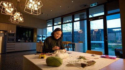 Person sewing a textile piece at a table with yarn and craft materials in a warmly lit community spa