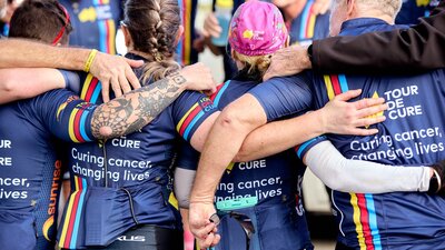 Signature Tour cyclists in kit, in a huddle with arms around each other,  taken from behind them