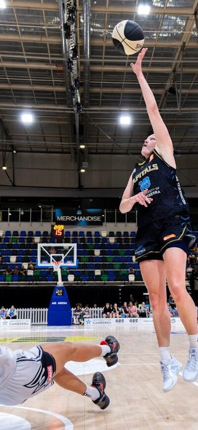 A UC Capitals player shooting a basketball towards the basket while the defender slides on the floor