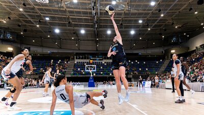 A UC Capitals player shooting a basketball towards the basket while the defender slides on the floor