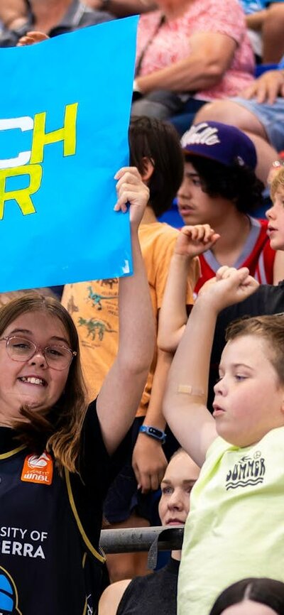 Two young fans cheering in the stands while waving a UC Capitals poster.