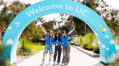 3 excited students beneath a decorative arch that reads "Welcome to UC"