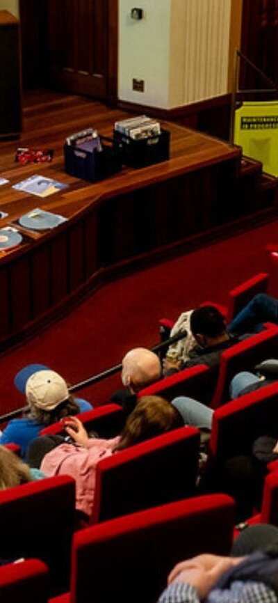 Audience in a theatre and a man at the front preparing to play a vinyl