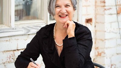 A smiling woman with grey hair sits at a table outdoors, writing in a notebook.